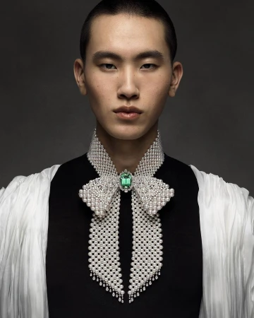 Close-up fashion shot of an Asian male model with a shaved head wearing a black top and large pearl-encrusted bow-tie necklace with a green jewel.