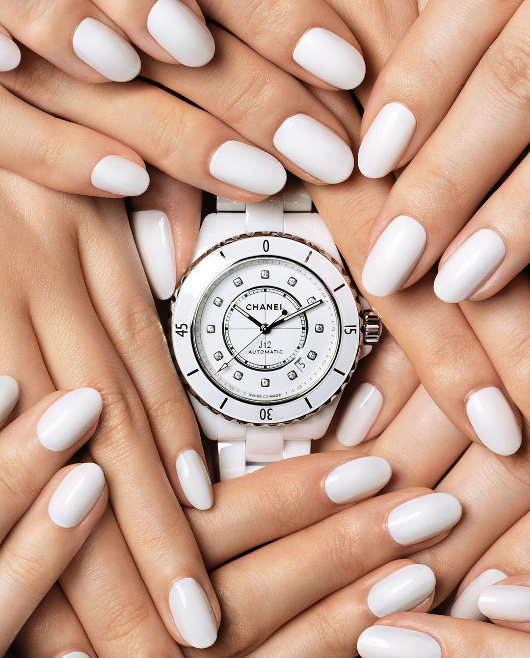 Close-up of several hands with white manicured nails surrounding a white ceramic Chanel watch, highlighting fashion and beauty.
