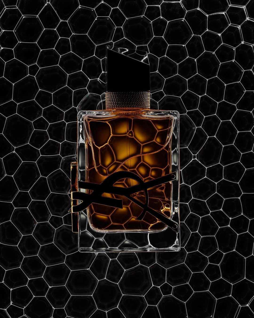 Studio shot of a fragrance bottle with black YSL logo, set against a geometric honeycomb backdrop. The bottle features brown liquid inside.