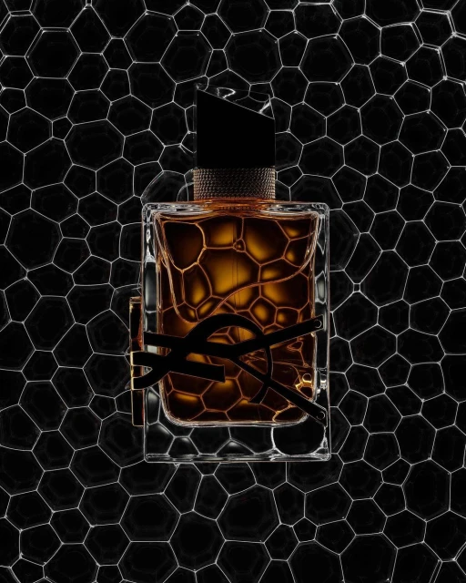 Studio shot of a fragrance bottle with black YSL logo, set against a geometric honeycomb backdrop. The bottle features brown liquid inside.