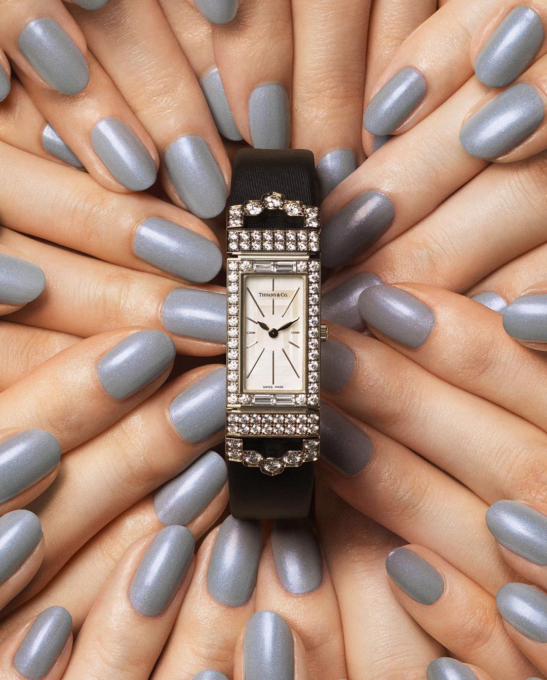 Close-up of a rectangular diamond watch surrounded by manicured hands with light gray nail polish, fashion accessories