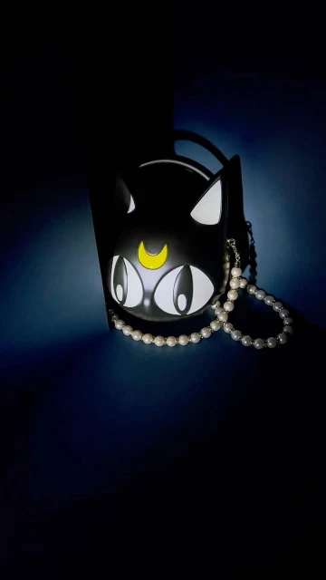 Cat-shaped black handbag with white trim, gold moon detail, and pearl strap in a low-light setting against a dark backdrop.