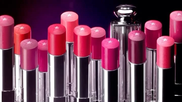 A row of lipsticks in various shades of pinks and reds, along with a perfume bottle, set against a gradient purple background.