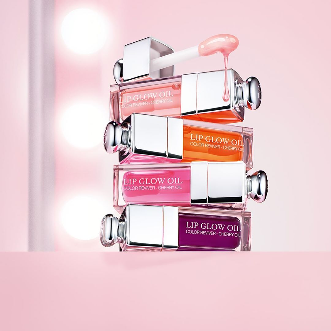 Stack of four 'Lip Glow Oil' lip glosses by Dior in light pink, orange, bright pink, and deep purple on a light pink surface.