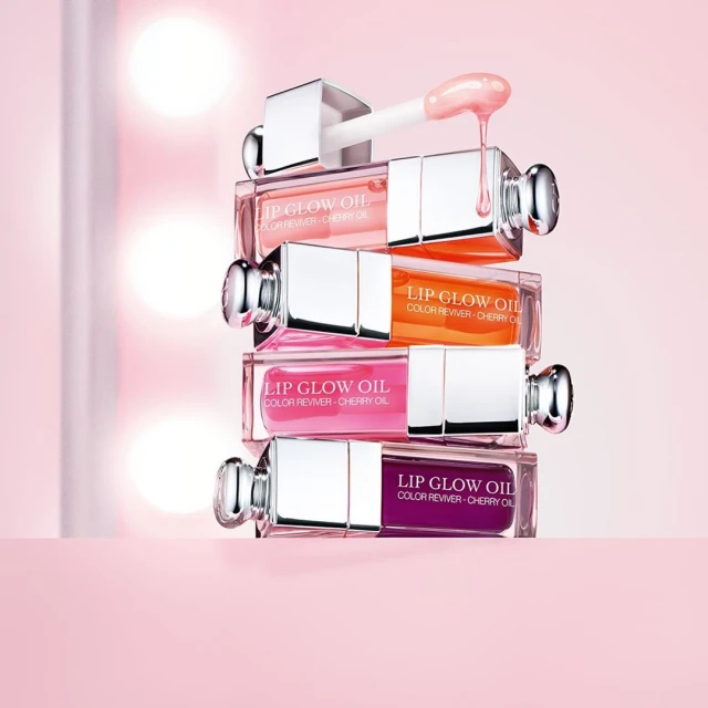 Stack of four 'Lip Glow Oil' lip glosses by Dior in light pink, orange, bright pink, and deep purple on a light pink surface.