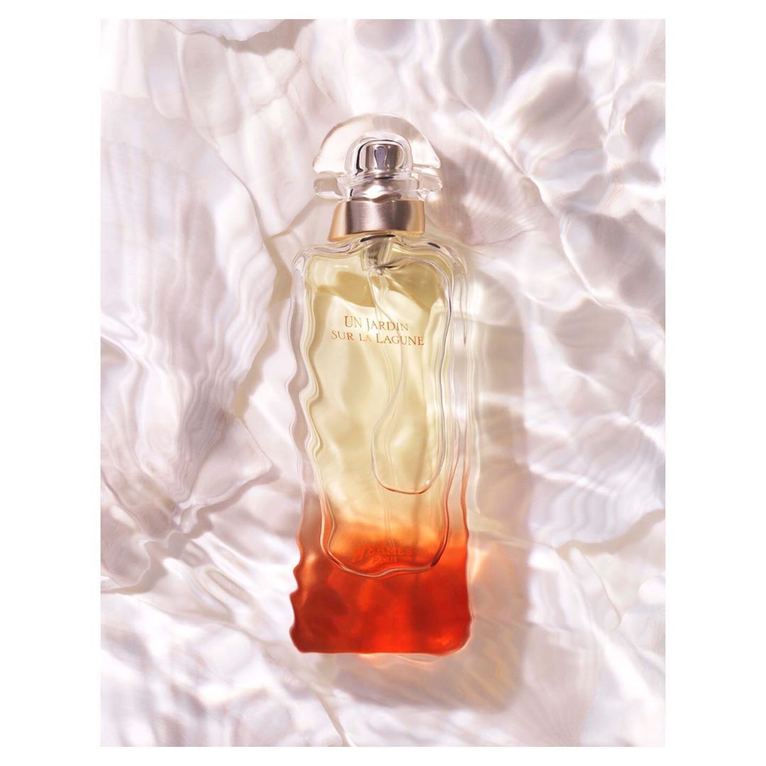 A perfume bottle by Hermès with a gradient fill is the primary subject, set against a white background with refracted light patterns.