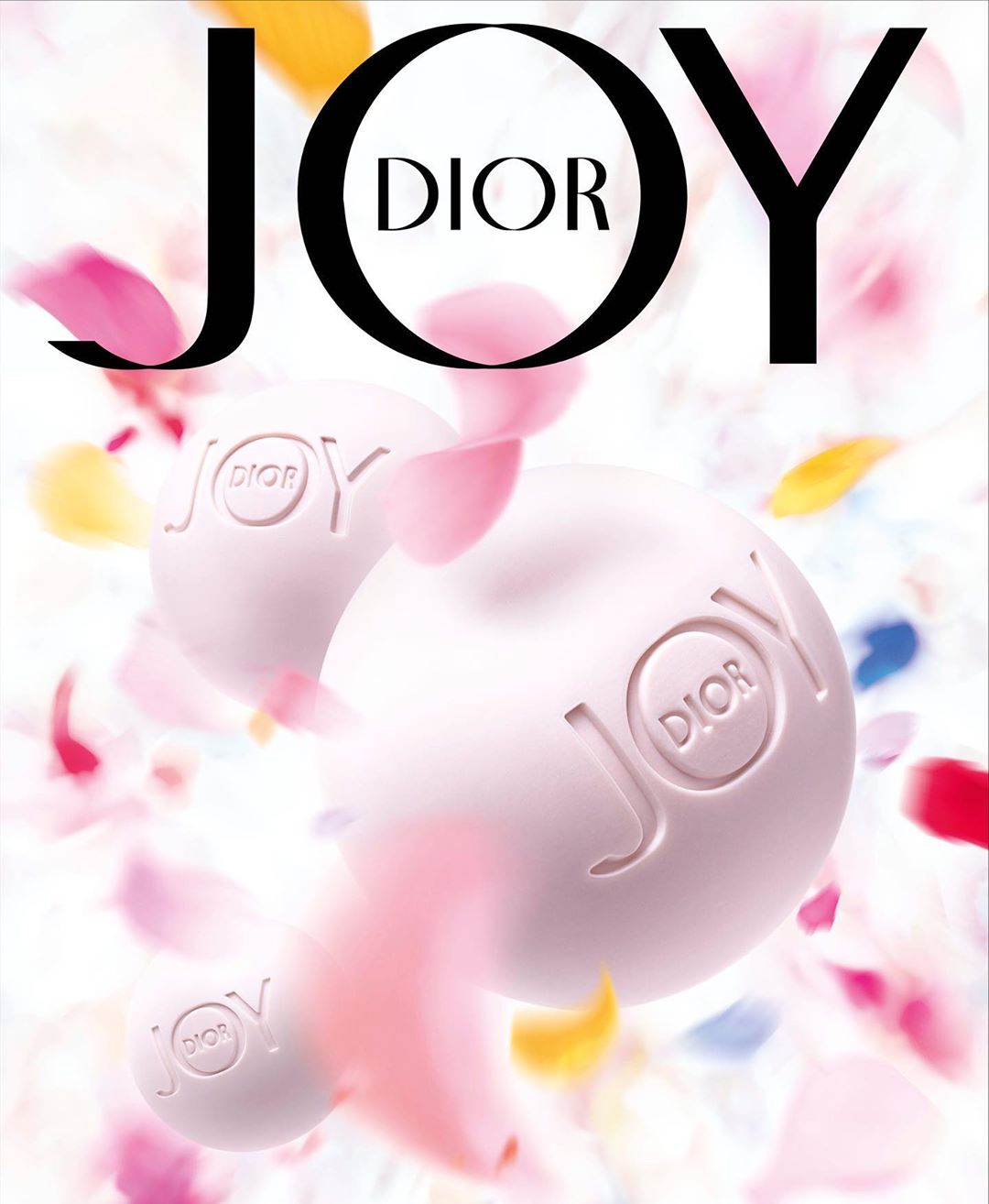 JOY by Dior advertisement featuring two perfume spheres with colorful flower petals falling around them on a white background.