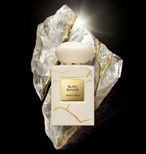 A bottle of Blanc Kogane Armani Prive perfume is displayed against a light-reflecting stone. Creamy white bottle with a rounded cap.