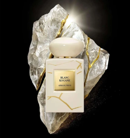 A bottle of Blanc Kogane Armani Prive perfume is displayed against a light-reflecting stone. Creamy white bottle with a rounded cap.