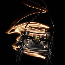 Transparent handbag with gold hardware and black fringe hanging against a dark background with golden light trails