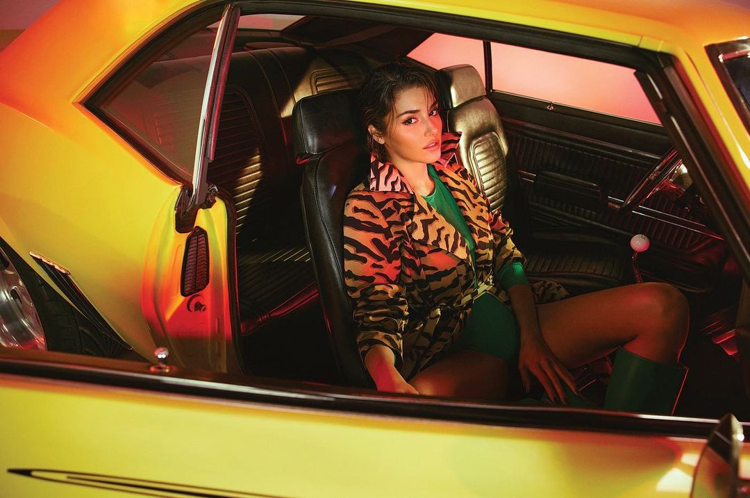 Stylish woman in tiger-print jacket poses inside a yellow classic car interior with leather seats and red-lit background. Automotive fashion.