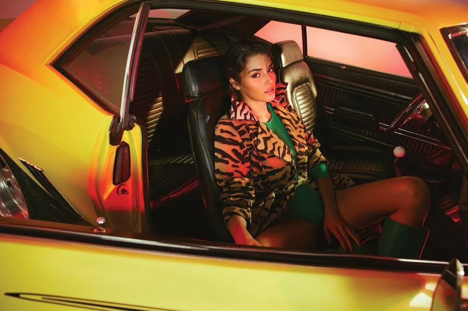 Stylish woman in tiger-print jacket poses inside a yellow classic car interior with leather seats and red-lit background. Automotive fashion.