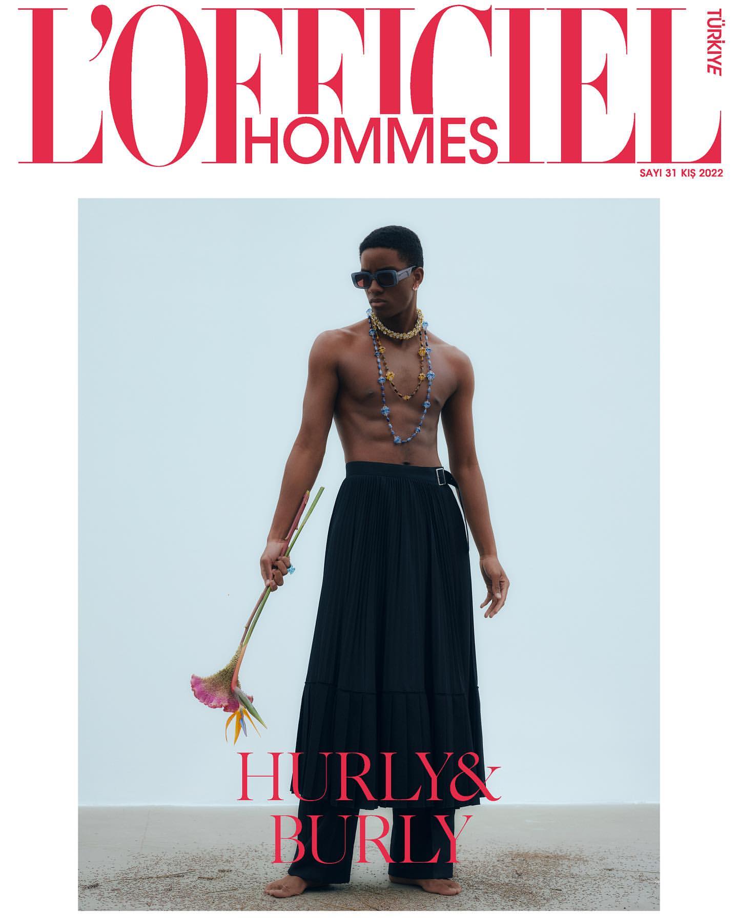 Fashion editorial cover featuring a shirtless, barefoot male model in a black skirt, holding a flower, with the L'Officiel Hommes masthead.
