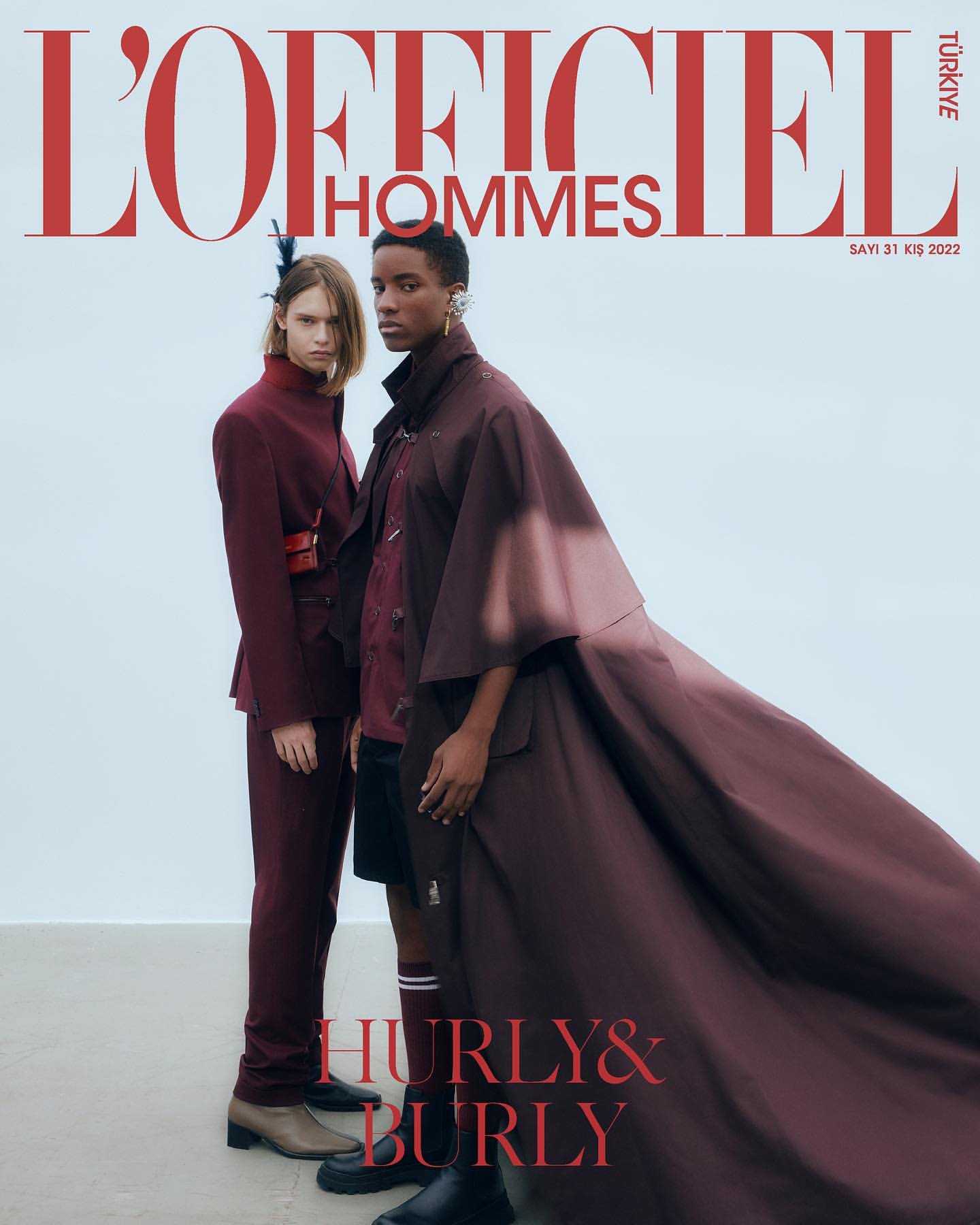 Two fashion models pose on a cover of L'Officiel Hommes magazine, wearing matching brown outfits. White backdrop with magazine title in red.