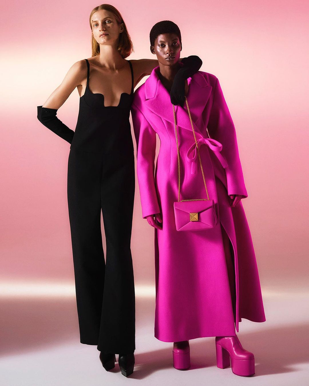 Two fashion models pose in front of a pink gradient background, one in a black jumpsuit and one in a bright pink coat and platforms.