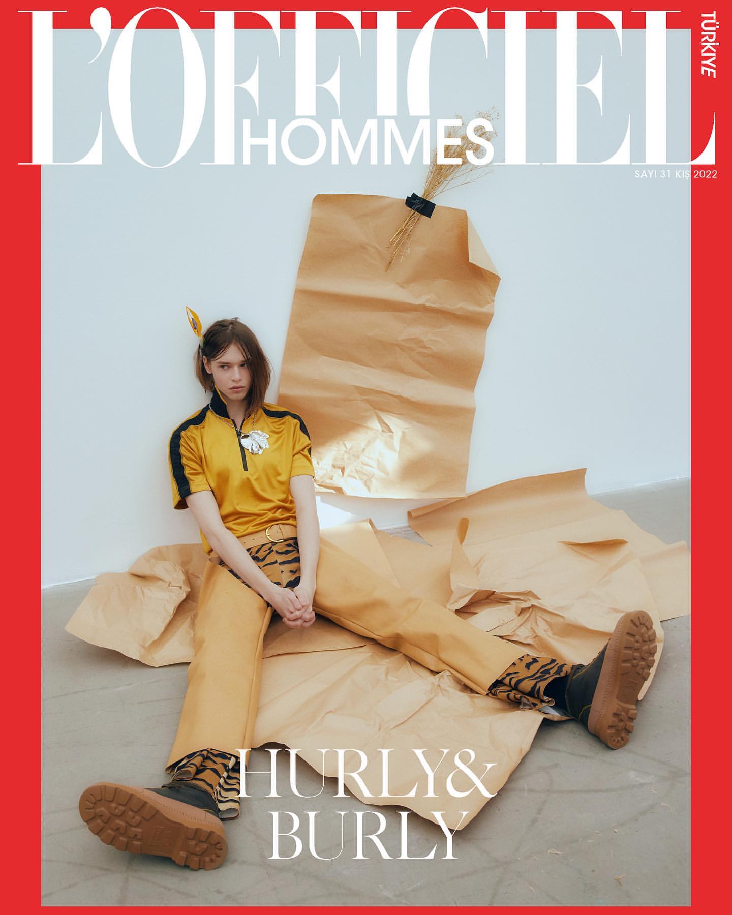 L'Officiel Hommes Turkey cover featuring a model in yellow shirt and pants, sitting on craft paper against a white backdrop and red border.