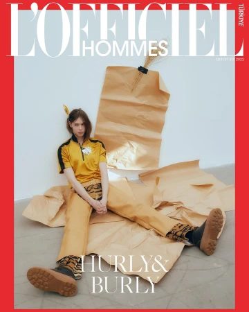 L'Officiel Hommes Turkey cover featuring a model in yellow shirt and pants, sitting on craft paper against a white backdrop and red border.