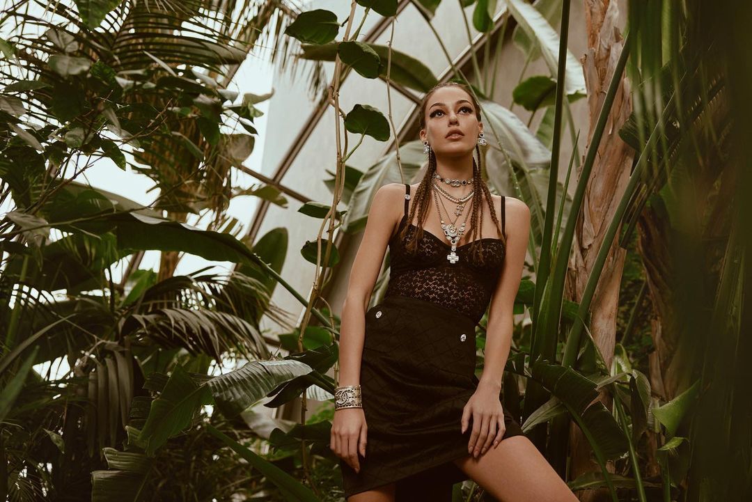 Woman in a black dress and jewelry posing in a verdant indoor garden with palm fronds and natural light coming through a glass ceiling.
