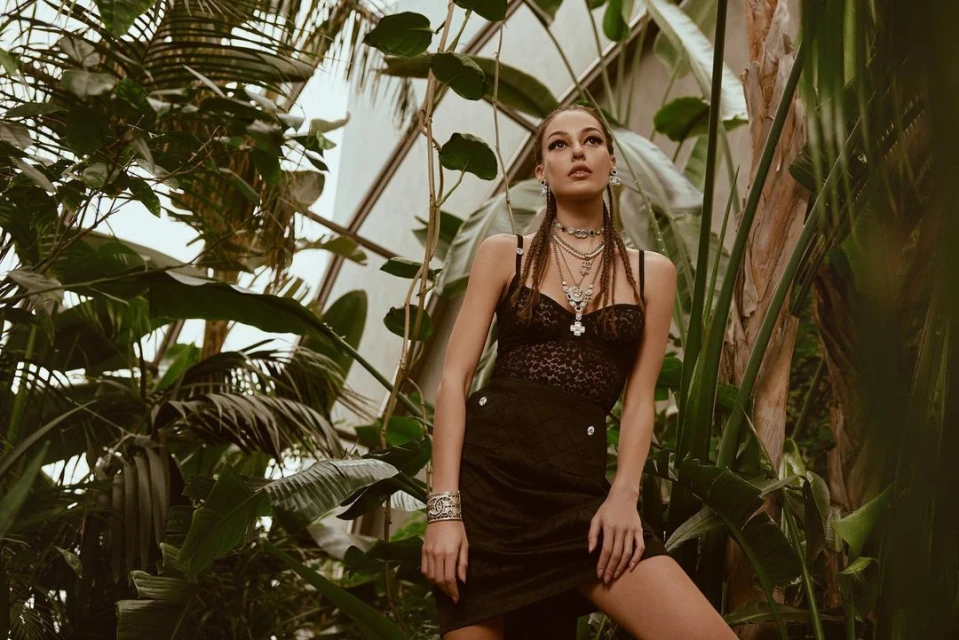 Woman in a black dress and jewelry posing in a verdant indoor garden with palm fronds and natural light coming through a glass ceiling.