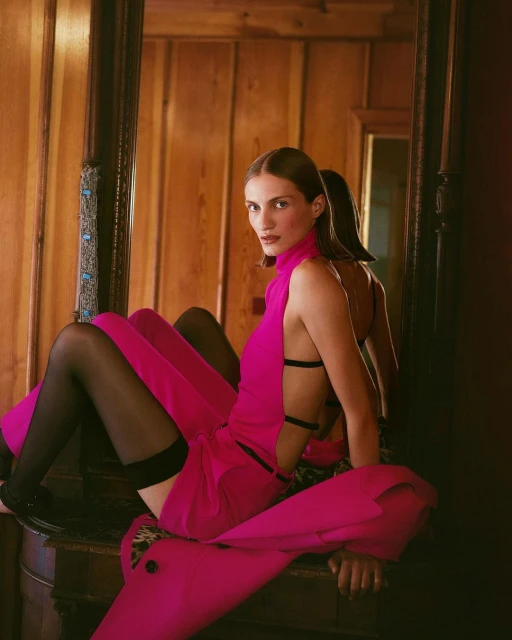 Elegant woman in a vibrant pink outfit poses in front of an antique mirror, reflecting a rustic wood-paneled room with warm lighting.