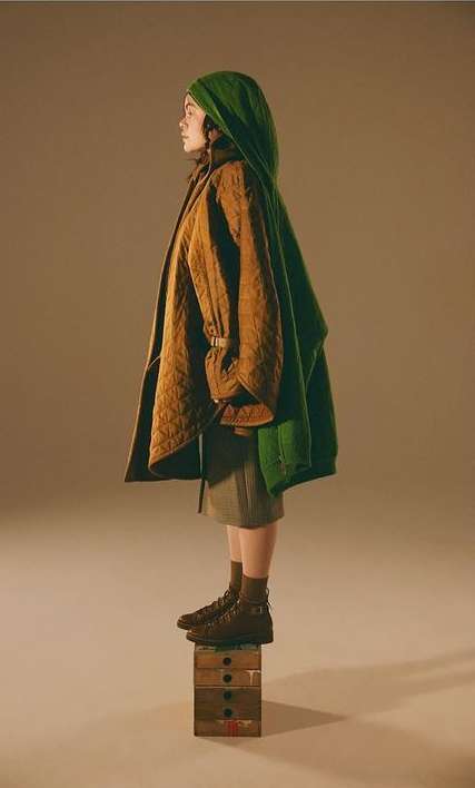 Side profile studio shot of adult female in brown boots, olive skirt, green head covering, and tan jacket standing on painted wooden box.