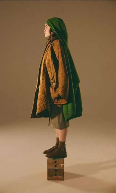 Side profile studio shot of adult female in brown boots, olive skirt, green head covering, and tan jacket standing on painted wooden box.