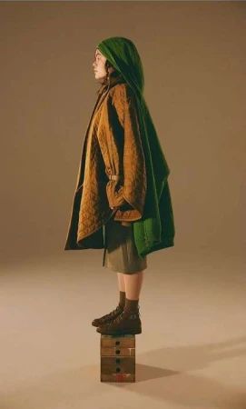 Side profile studio shot of adult female in brown boots, olive skirt, green head covering, and tan jacket standing on painted wooden box.