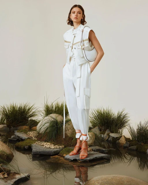 A woman models white denim vest, pants, purse, and strappy sandals in a studio with decorative rocks, moss, and small pools of water.