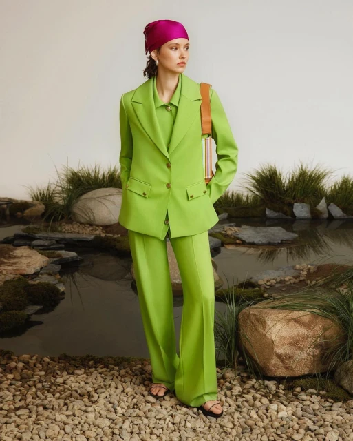 A stylish adult female in a green suit and pink headscarf stands outdoors by water with rocks and plants for a fashion shoot.