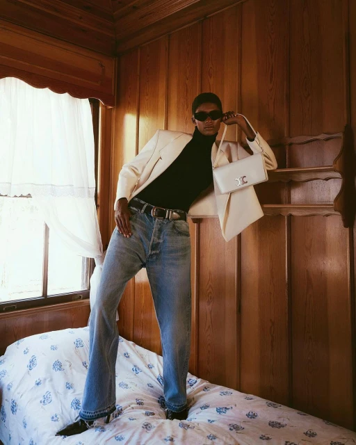 Fashion shot of a woman with dark skin wearing jeans and a white jacket standing on a bed with a white purse in an indoor setting.