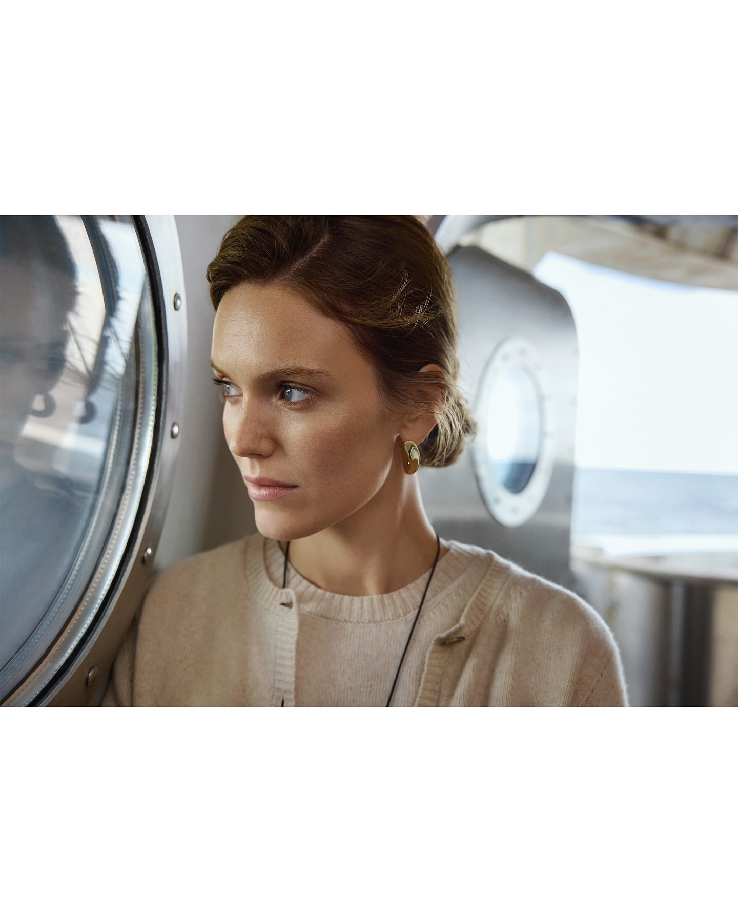 Portrait of an adult woman with short brown hair, wearing a cardigan sweater and earrings, standing near a metal structure.