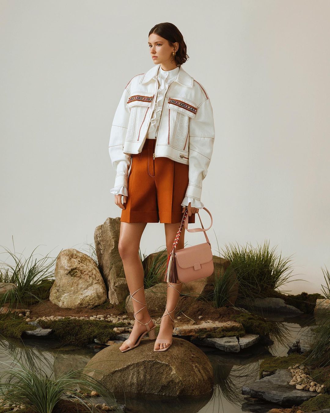 Model standing on a mossy rock in an indoor water set wearing a white patterned jacket, rust shorts, and holding a pink handbag