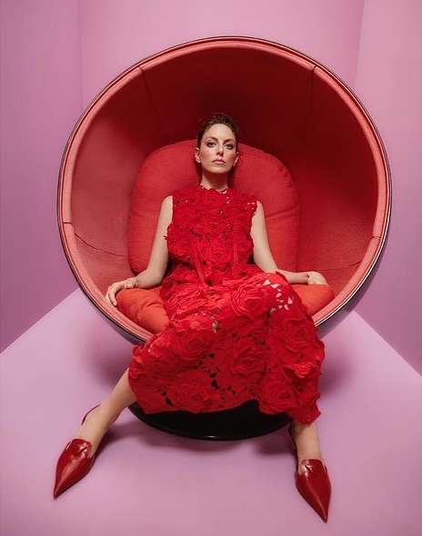 A woman in a red lace dress sits in a red egg chair against a pink wall, creating a striking monochromatic fashion portrait.