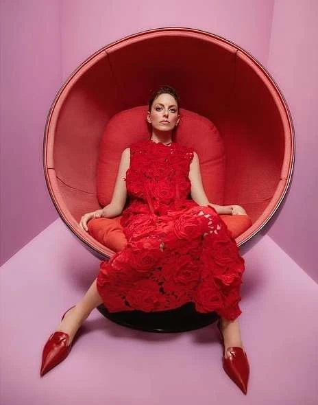 A woman in a red lace dress sits in a red egg chair against a pink wall, creating a striking monochromatic fashion portrait.