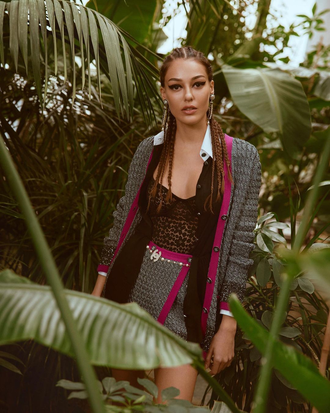 Adult female poses among lush greenery in a grey tweed skirt suit with pink accents, complemented by delicate earrings and braided hair.