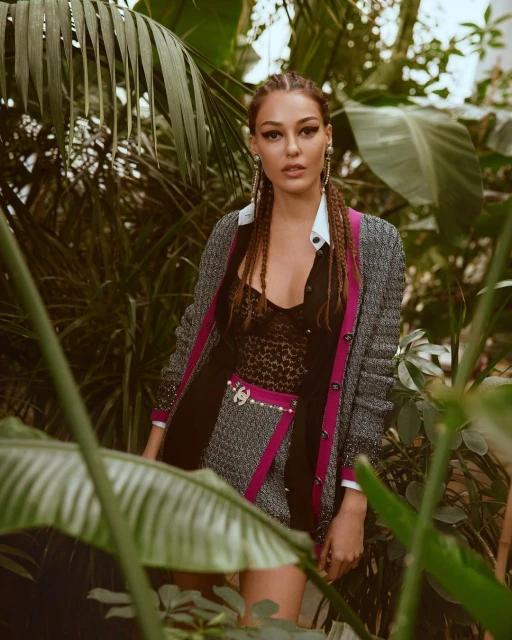 Adult female poses among lush greenery in a grey tweed skirt suit with pink accents, complemented by delicate earrings and braided hair.