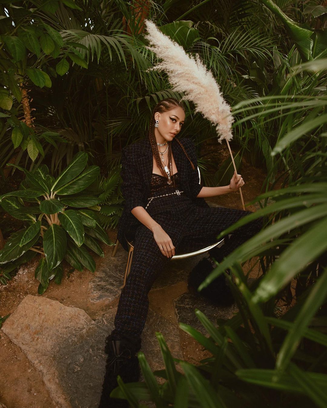 Woman in black suit poses with ornamental grass among lush greenery, creating an exotic fashion editorial look in a natural environment.