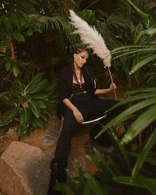 Woman in black suit poses with ornamental grass among lush greenery, creating an exotic fashion editorial look in a natural environment.