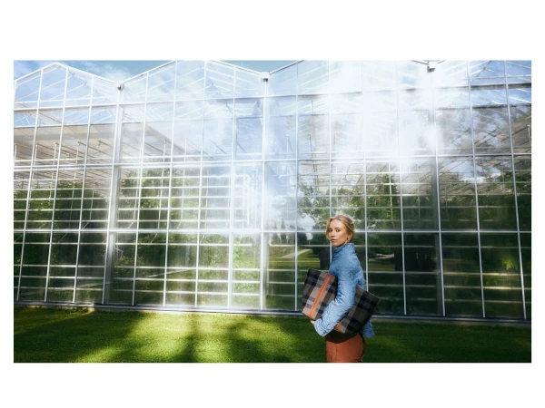 Woman carrying a plaid tote bag outdoors in front of a large modern glass greenhouse structure