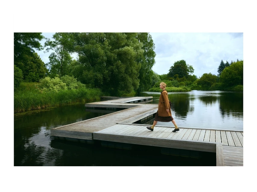 Adult woman in a coat walks on a wooden platform over a lake surrounded by greenery. Nature and architecture combine in this outdoor scene.