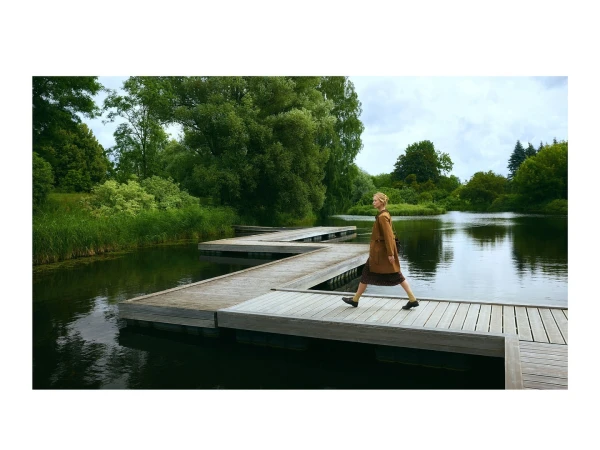 Adult woman in a coat walks on a wooden platform over a lake surrounded by greenery. Nature and architecture combine in this outdoor scene.