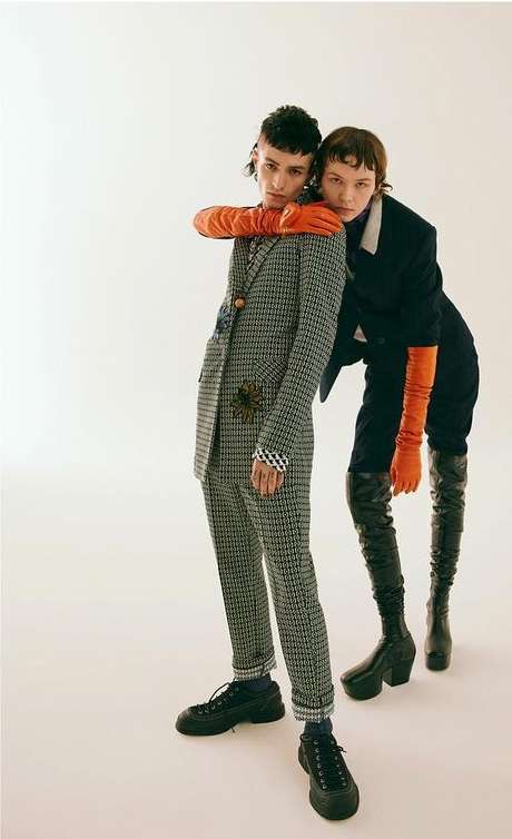 Two models in a studio setting. One is wearing a grey suit and black sneakers while the other wears a black suit with knee-high black boots.