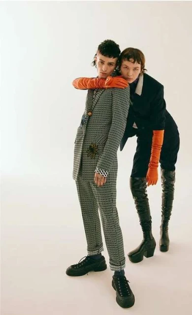 Two models in a studio setting. One is wearing a grey suit and black sneakers while the other wears a black suit with knee-high black boots.