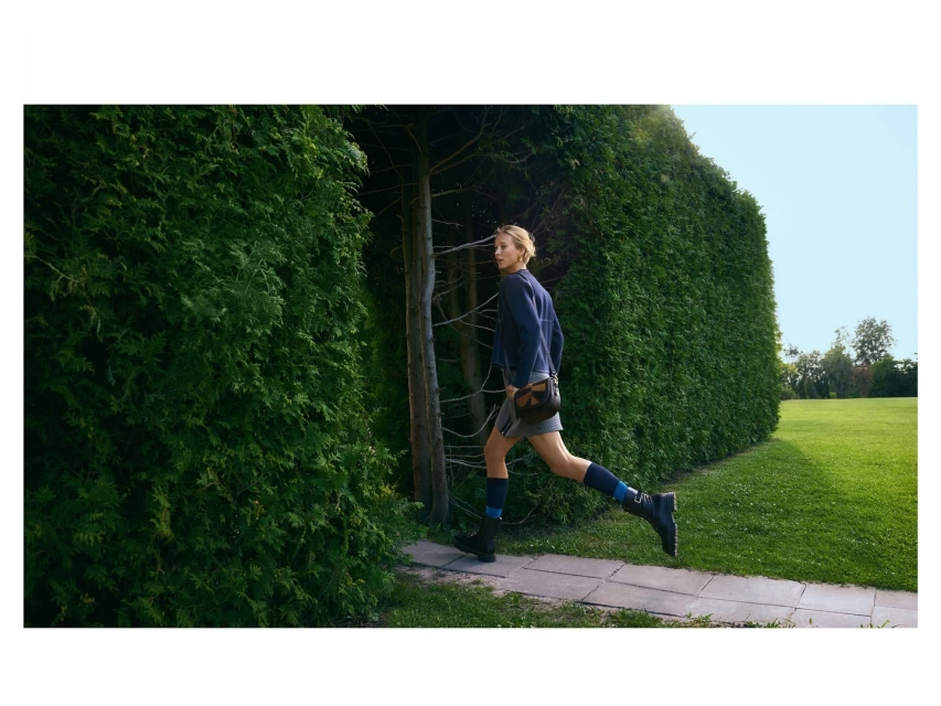 Young woman in a blue jacket and grey skirt runs through a hedge opening into a green lawn with trees under a blue sky, outdoors.