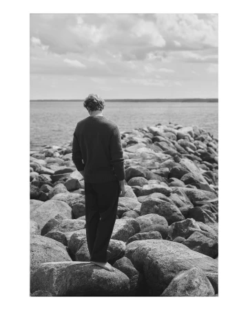 Monochrome photograph of a man standing on a rocky jetty overlooking a large body of water under a cloudy sky.