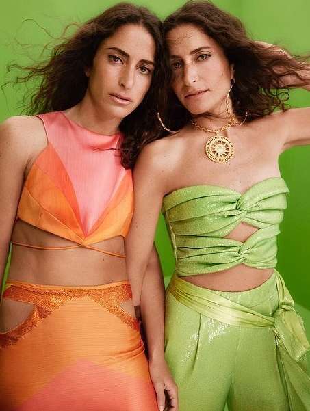 Two adult females pose against a green background, wearing orange and green shiny textured outfits, gold jewelry, and wavy hair. Portrait shot.