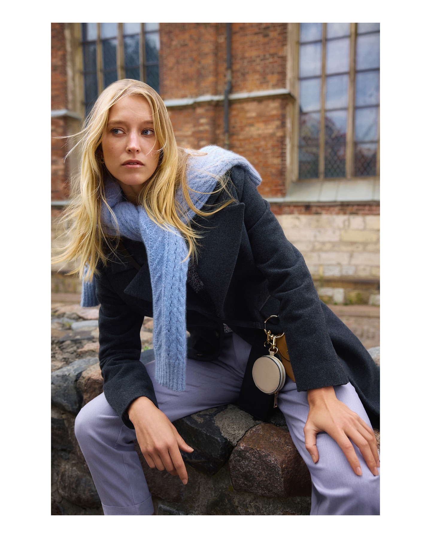 A blonde woman sits on a stone wall outdoors. She wears a dark coat, light purple pants, and a light blue scarf. A building is visible behind her.