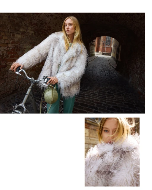 Fashion shot of a woman in a furry coat posing with a bicycle in a brick alleyway with a purse; indoor lighting, medium shot.