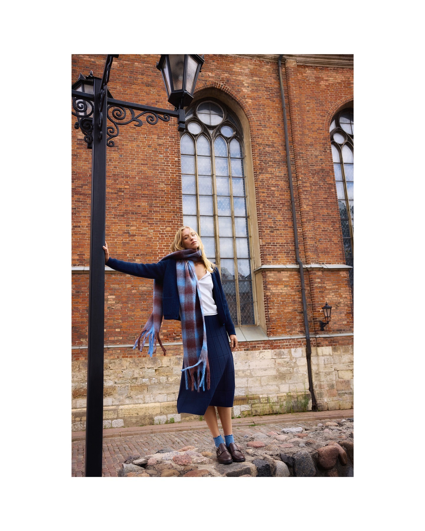 A blonde woman in a navy skirt and blazer poses next to a black street lamp and brick building in an urban environment, wearing a plaid scarf.
