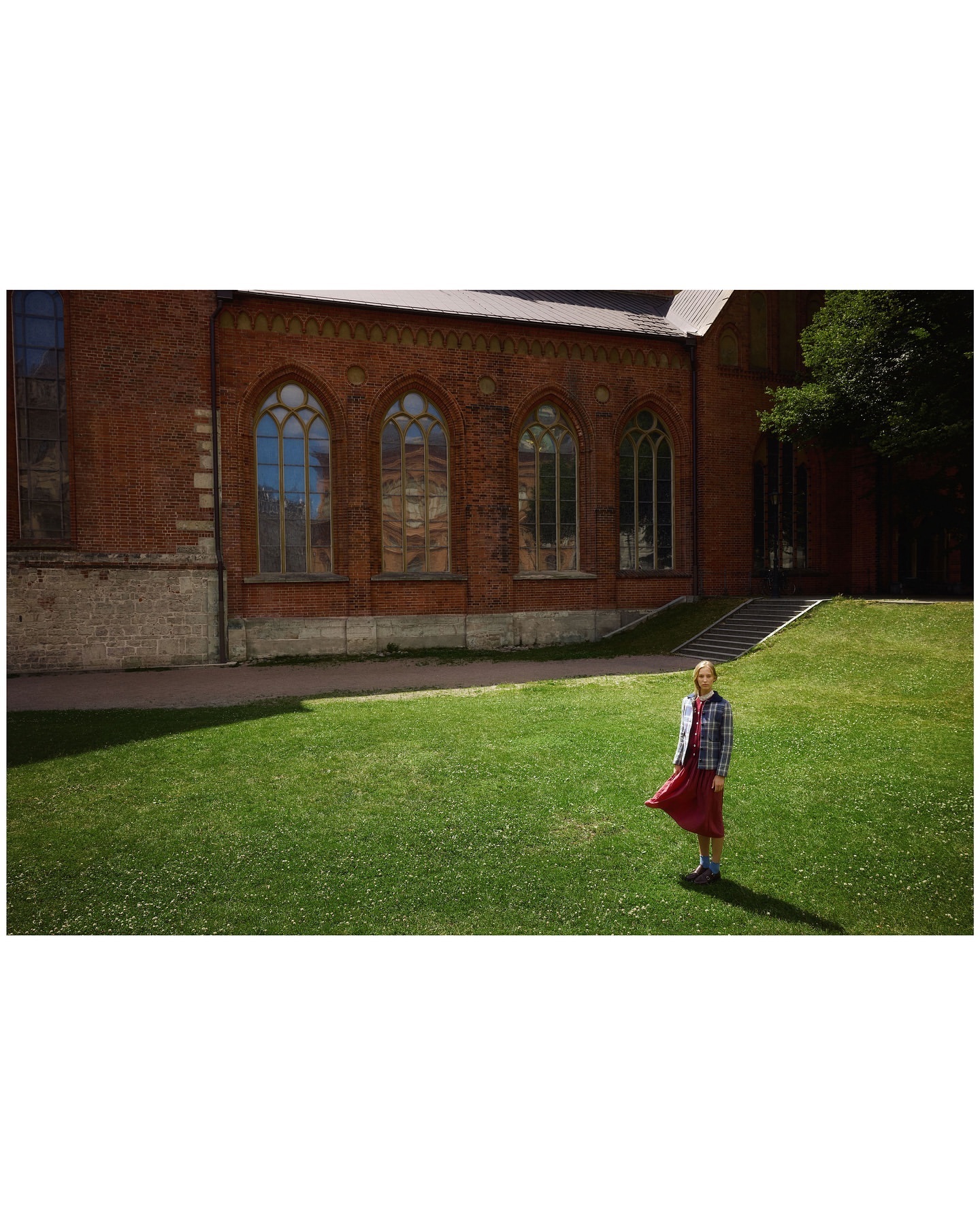 Young woman stands on the green lawn outside the red brick building, wearing a plaid shirt and long red skirt, on a sunny day.